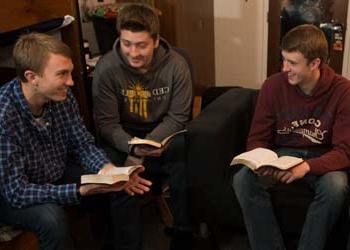 Three male students gather in a softly lit dorm room to study the Bible